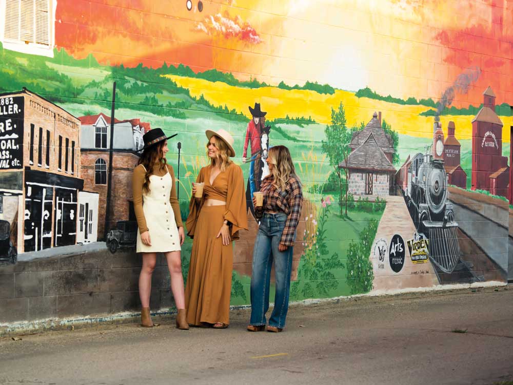 Mural on the side of a building in Virden, Manitoba showing historic buildings, train locomotive, elevators. Three women stand in front of the mural.