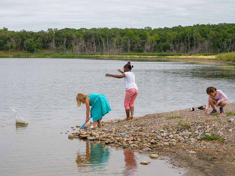 Three girls play on the rocky shore of Salt Lake, Manitoba. The small lake is surround by trees.